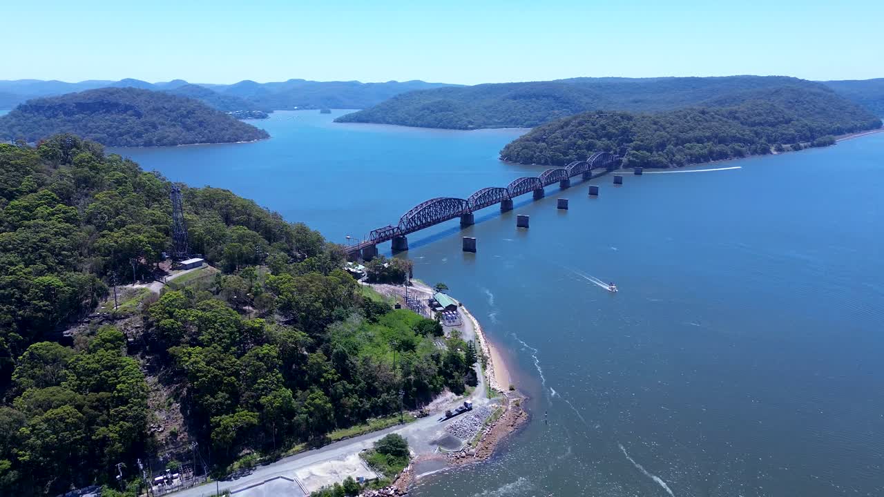 Paesaggio aereo con drone di una barca in navigazione vicino al deposito di Long Island con alberi della boscaglia lungo la costa del promontorio e il ponte ferroviario del fiume Hawkesbury sullo sfondo. Brooklyn, Australia, viaggi, turismo.