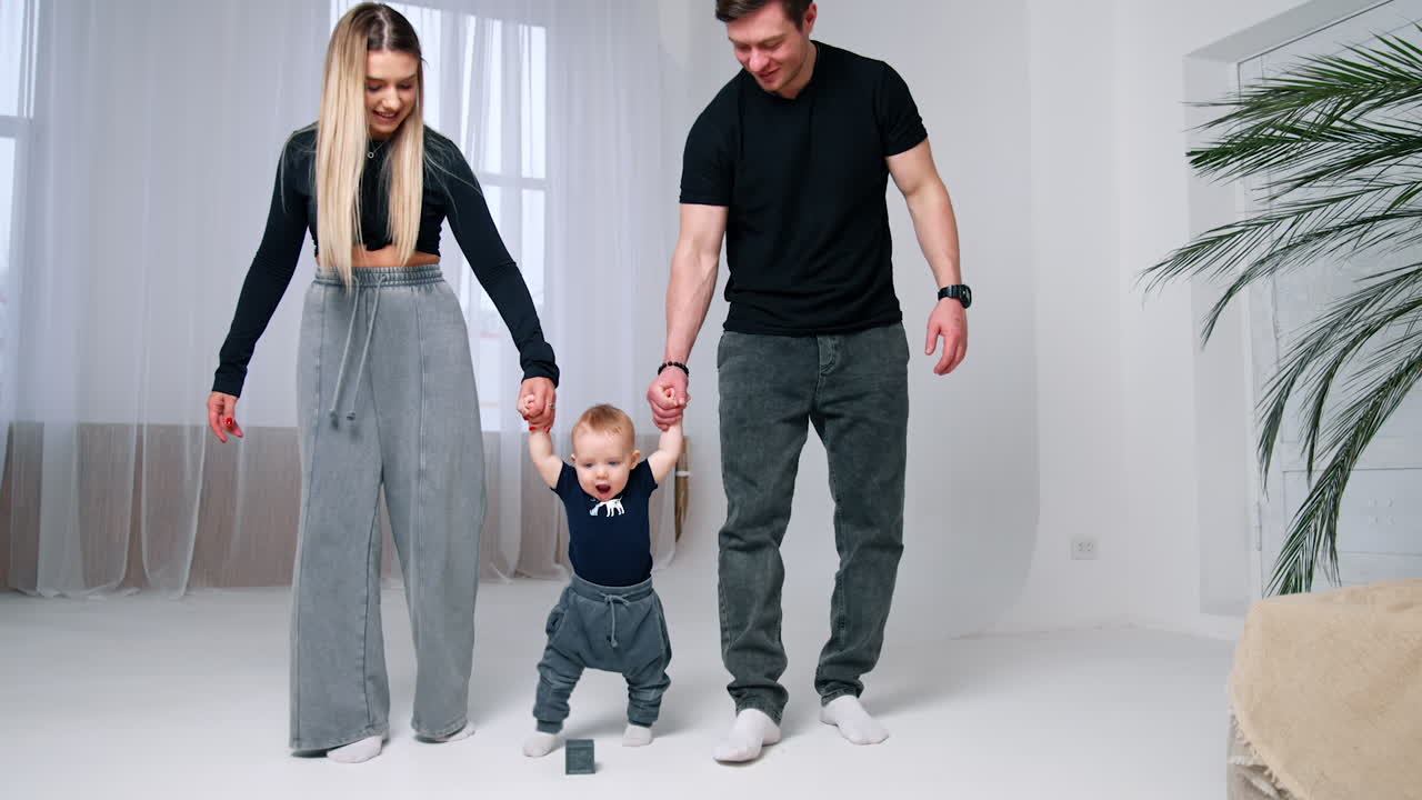 Adorable Caucasian baby boy walks kicking the cube. Parents hold their child by the hands.