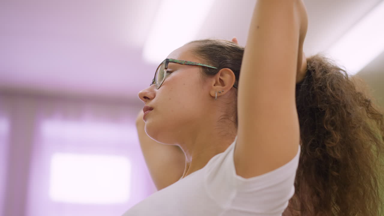 Close view woman in white top and glasses arranging hair in blurred studio background, hands styling strands, calm expression, light filled soft portrait capturing motion and texture
