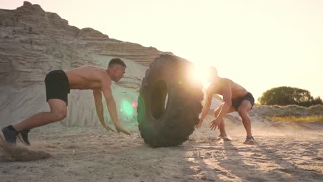 two muscular open-chested athletes train in active mode on the beach doing push-ups and pushing a huge wheel against a sandy mountain at sunset