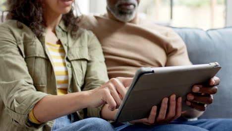una pareja biracial feliz sentada en el sofá y usando una tableta en casa, en cámara lenta