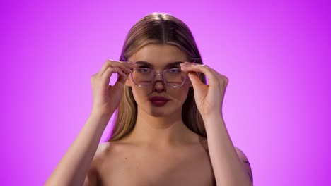 medium shot of a young pretty woman who needs a visual aid because her eyesight is not good enough and puts on glasses and looks to the camera in front of pink background in slow motion