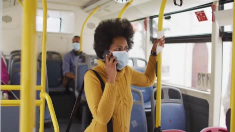 Afroamerikanische-Geschäftsfrau-Mit-Gesichtsmaske,-Die-Auf-Dem-Smartphone-Spricht-Und-Im-Bus-Steht