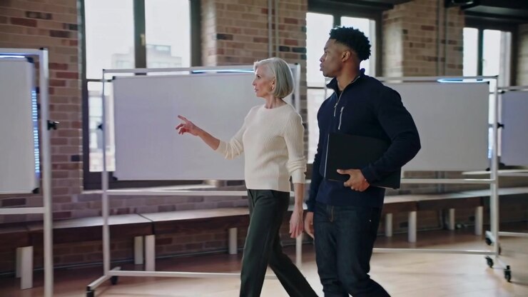 Entering left, senior woman sweater leading, man hoodie with laptop surveying whiteboards in loft