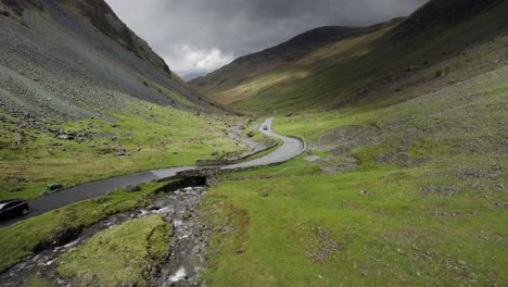 A-car-drives-over-a-an-old-stone-bridge-in-a-valley,-Honister-pass-towards-a-slate-mine-in-the-English-Lake-District-on-a-moody-overcast-day