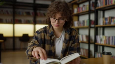 confident girl student with curly hair in glasses and a plaid shirt reads a book while sitting at a table in the library