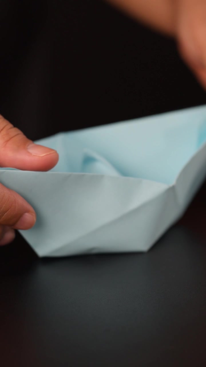 Close-up hands carefully folding light blue origami paper boat on dark tabletop, shallow focus