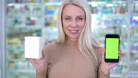 gorgeous confident caucasian woman posing with chromakey smartphone and pills box in pharmacy. portrait of beautiful blond female client looking at camera smiling advertising drugstore service.