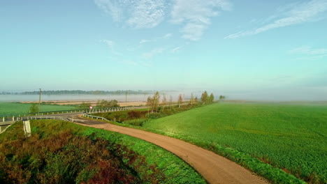 Ascending-drone-shot-over-misty-rural-fields-and-road-in-countryside-during-autumn-and-blue-sky