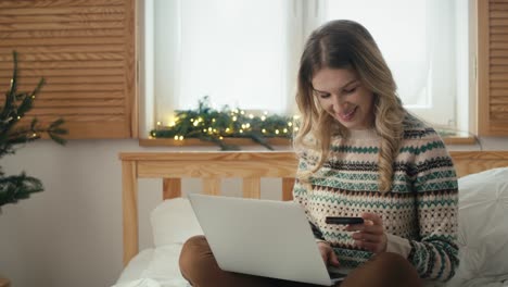 Cheerful-caucasian-woman-sitting-on-bed-and-doing-Christmas-shopping-with-use-of-laptop-and-credit-card