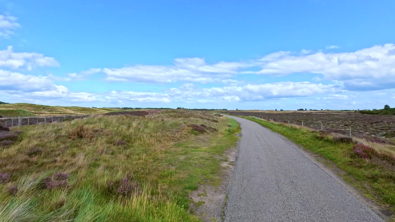 A vehicle travels down a narrow, winding country road through open moorland in Cromarty, Scotland, under bright daylight with steady forward camera movement