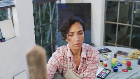 biracial female artist in apron holding brush and looking at canvas in studio, slow motion