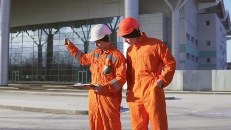 Two-construction-workers-in-orange-uniform-and-helmets-looking-over-plans-together.-Building-at-the-background