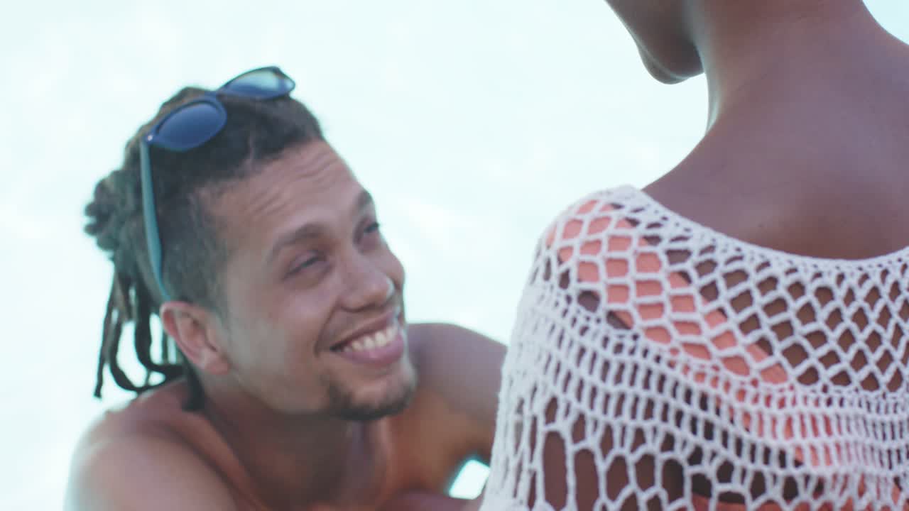 Casal na praia, a mulher virando-se e fazendo o homem inclinar-se, sorrindo, com saída de praia de crochê, espaço para texto.