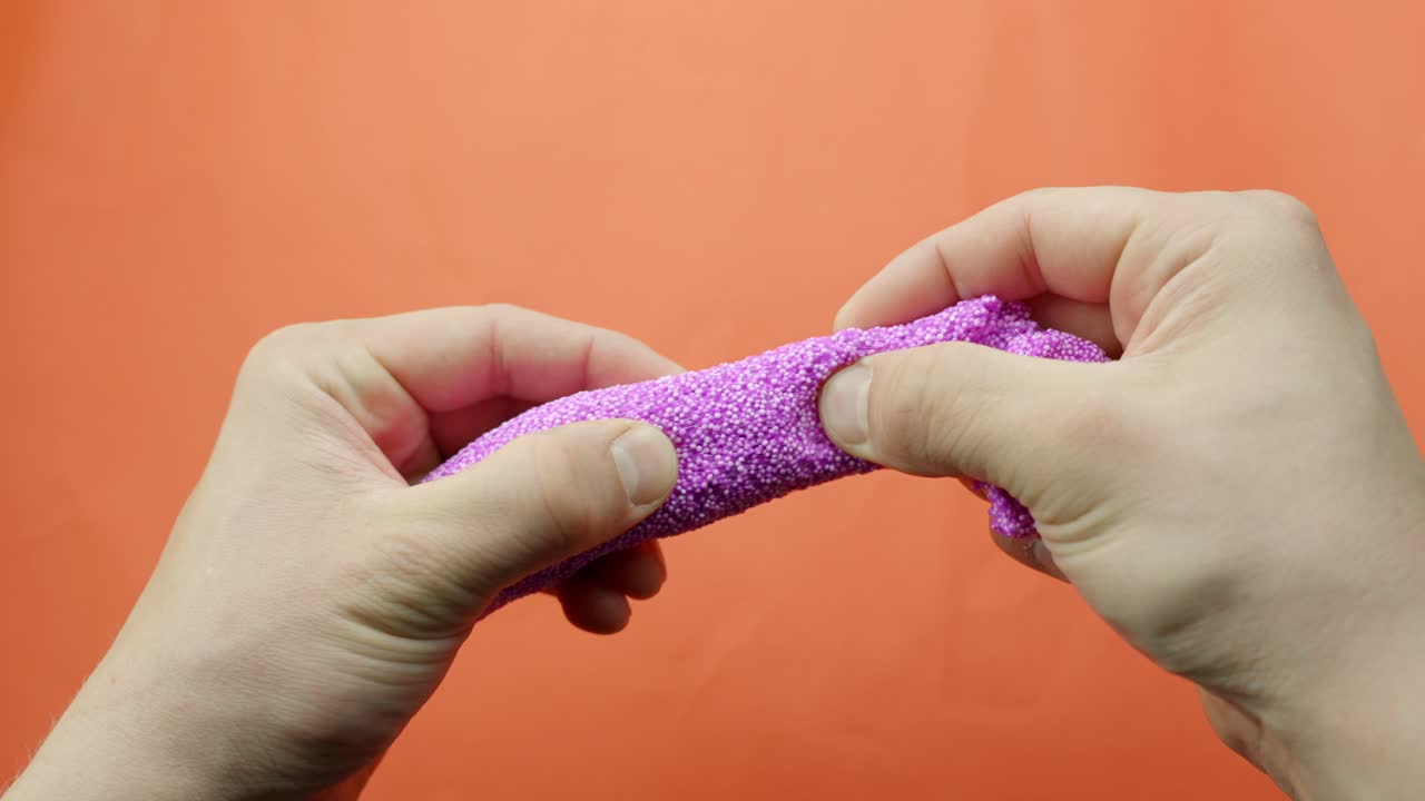 Cclose-up shows hands pulling and separating lilac-colored floam slime with foam beads for ASMR sensory play, revealing texture and stretch against orange backdrop, static camera