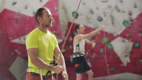 portrait of beautiful man rock climber belaying another climber with rope. indoors artificial climbing wall and equipment.