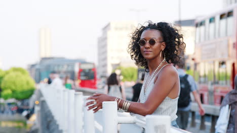 Fashionable-young-black-woman-leaning-on-the-handrail-of-a-bridge-in-the-city-looking-around,-side-view