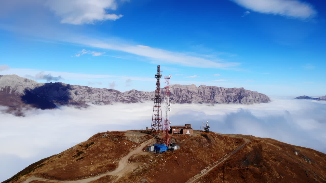 Trasmettitore in vetta alla montagna sopra le nuvole, drone aereo rivela una torre di telecomunicazioni color ruggine su un crinale brullo.