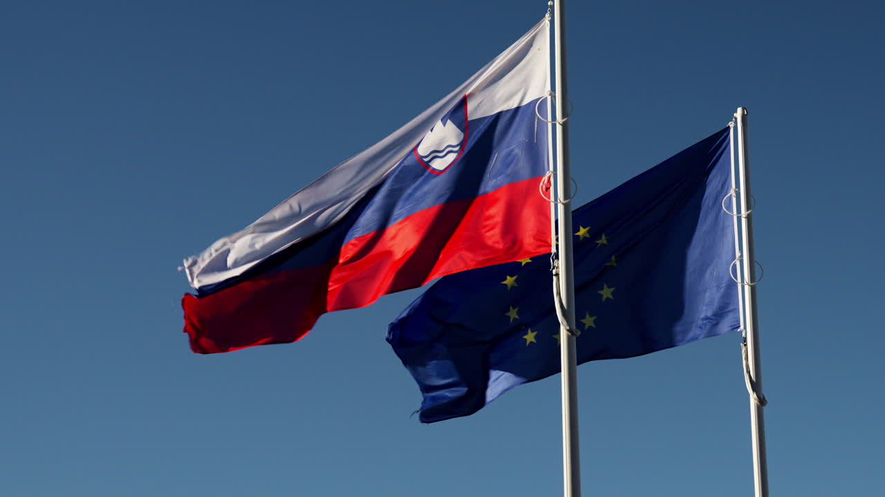 Açık güneşli bir gökyüzü altında Slovenya ve Avrupa bayrakları dalgalanıyor. Çekim görüntüsü, parlak gün ışığında farklı açılardan çekilmiş, hareket halindeki ulusal ve AB sembollerini gösteriyor.