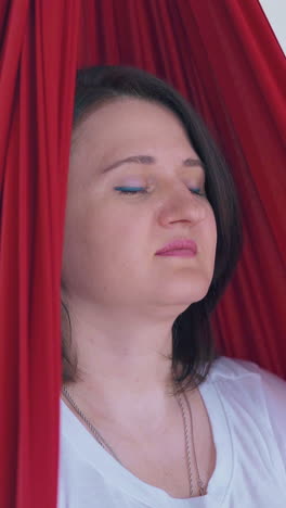calm woman in white t-shirt swings and relaxes in oriental fly yoga hammock meditating with closed eyes extreme closeup