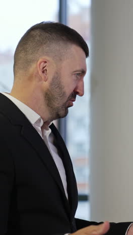 boss standing with his side and telling something to colleagues. male in suit explaining work points and gesturing with his hands. man in a big office. vertical video
