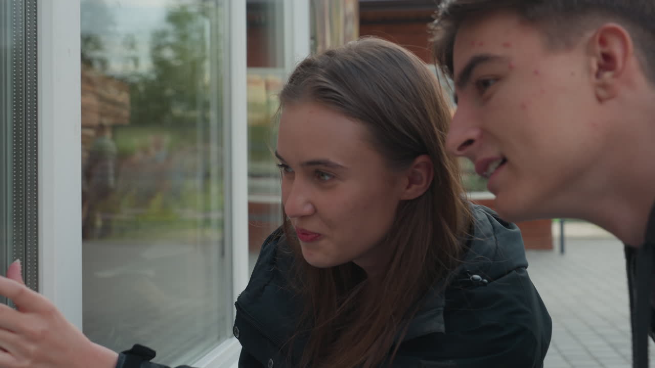 Amici adolescenti bianchi sorridenti alla finestra con interazione ravvicinata e risate calorose, atmosfera di prove spontanea con luce diurna soffusa, sfondo suburbano e zaini, intesa autentica come aspiranti attori