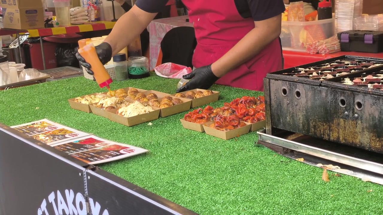 Ein Straßenhändler bereitet sorgfältig frische Takoyaki auf einer heißen Grillplatte zu. Die herzhaften Oktopusbällchen werden gewendet, bis sie goldbraun sind. Dieser beliebte Snack bietet einen köstlichen Geschmack des urbanen Streetfoods.