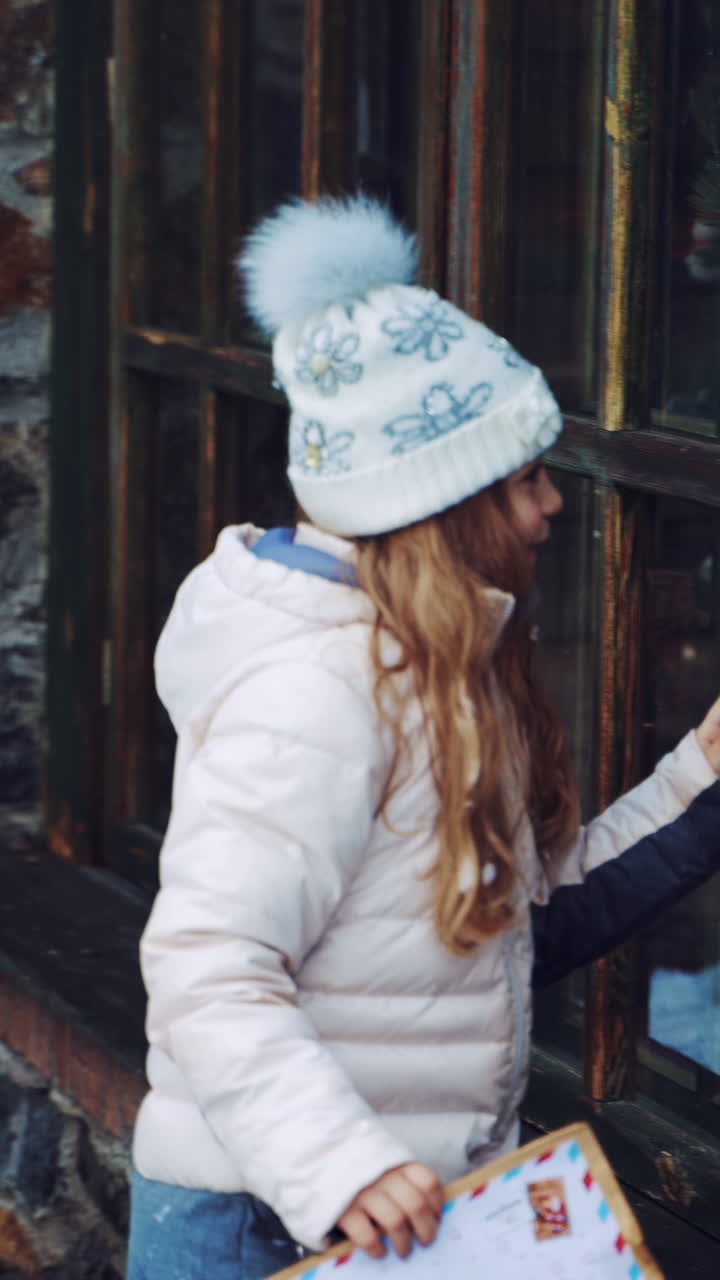 Little girl is looking through the small wooden windows outdoors. Curious child looks inside the house with fairy atmosphere while standing near the windows in winter. Vertical video