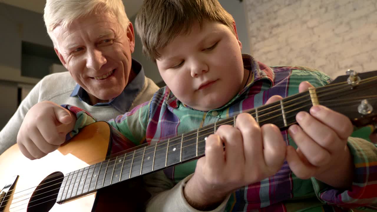Farfar lär sitt barnbarn att spela gitarr. En äldre man lär ett ungt barn att spela gitarr. Hemtrevnad, familjeidyll, mysighetskoncept, generationsskillnader. 60 fps