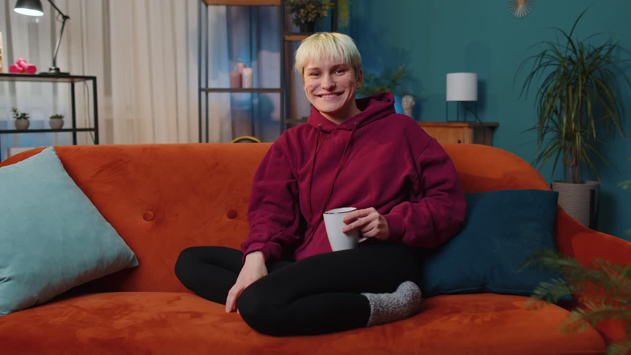 카메라에 친절하게 웃는 젊은 여성, 손을 흔들며 안 ⁇ 하세요, 안 ⁇ 하세요, 소파에서 집에서 인사합니다.