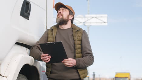 worker wearing vest and cap reading documents in a logistics park while is leaning on a truck 1