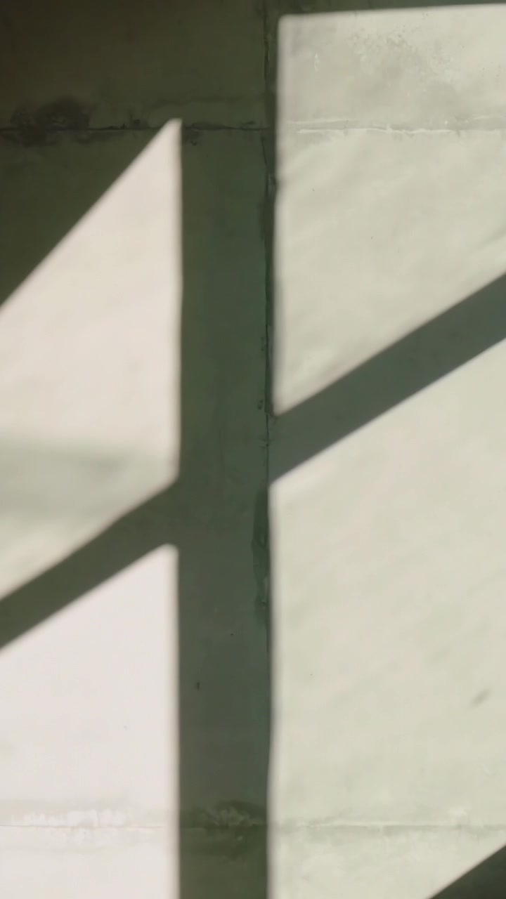 Vertical video: Casting sunlight via window making angular shadows shifting across pale wall seam