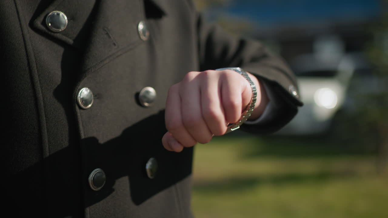 Close-up of black coat with metallic buttons as individual lowers hand after checking wristwatch, outdoor urban setting with blurred background features modern buildings, parked cars, greenery