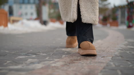 close-up view of girl wearing warm beige winter coat and brown suede boots walking on snowy path, textured sole of boots visible as she steps forward on cold pavement with snow-covered surroundings