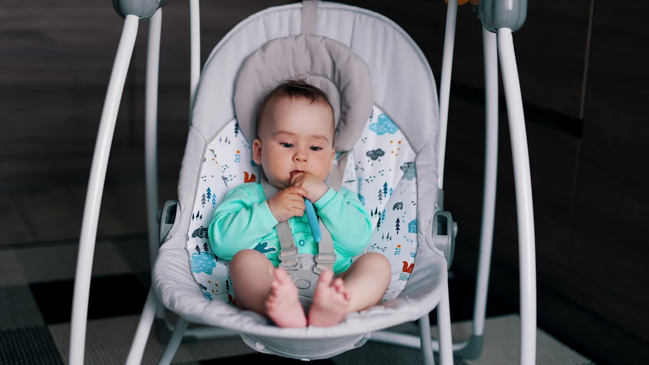 Beautiful child sitting in a swaying chair chews the spoon. Active healthy baby after feeding.