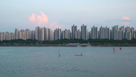 gente haciendo windsurf en el río han en jamshil rascacielos apartamento paisaje urbano en el fondo al atardecer, hombre en lancha tomando fotos de dos chicas en paddleboards en medio del río cerca de ttukseom