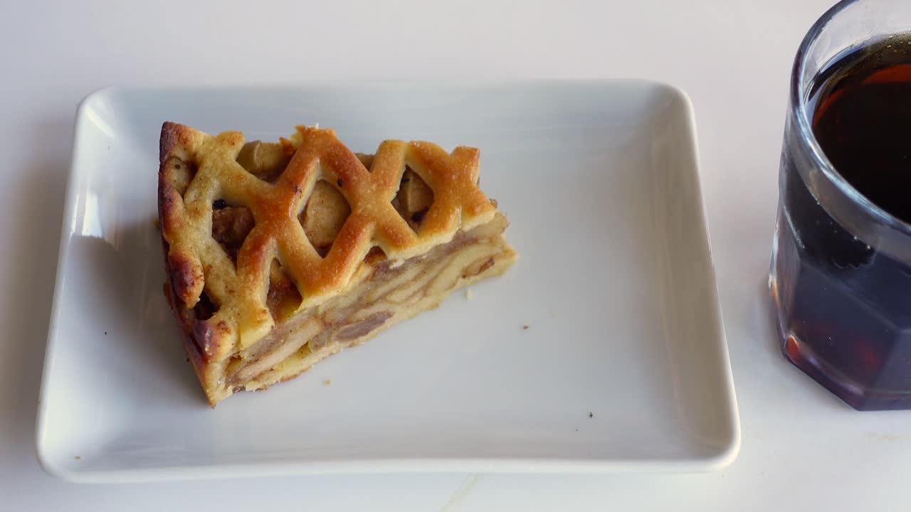 una rebanada de pastel de manzana con un tenedor en un plato blanco, y un vaso de bebida oscura
