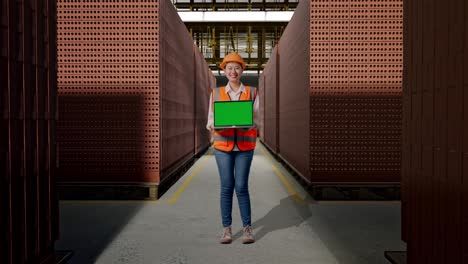 full body of asian female engineer with safety helmet smiling and showing green screen laptop to the camera with red brick packed in stacks are stored