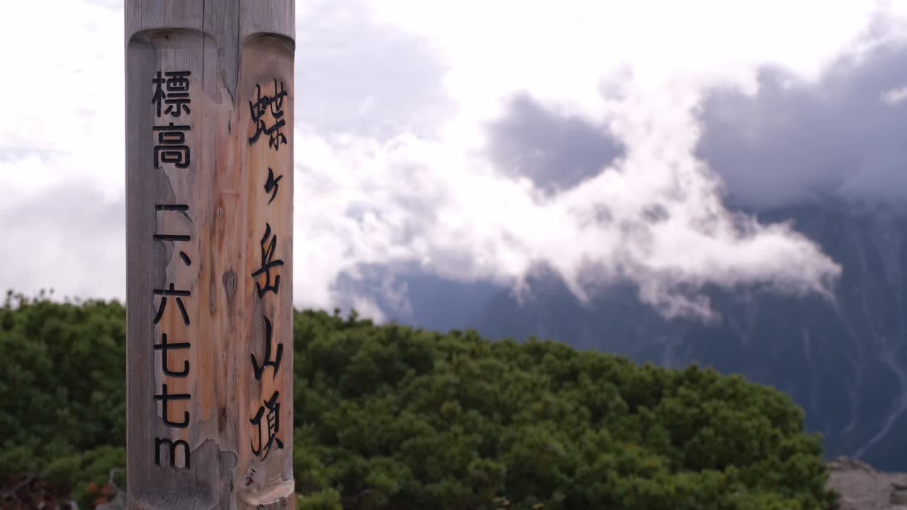 Close-up of the wooden Chogatake Summit post reading “Elevation 2677m,” with clouds drifting over the mountains behind it