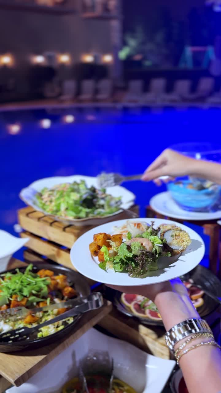 Close-up of a guest serving fresh green lettuce salad onto a plate with tongs. Healthy vegetable option at a night resort buffet dinner under blue lighting