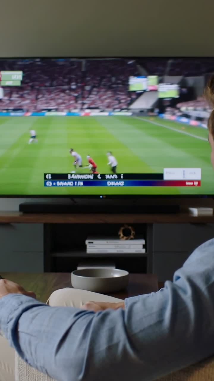 Video verticale: Uomo in camicia blu che reagisce all'aggiornamento del punteggio TV, spostando il telecomando a casa, con spazio per il testo.