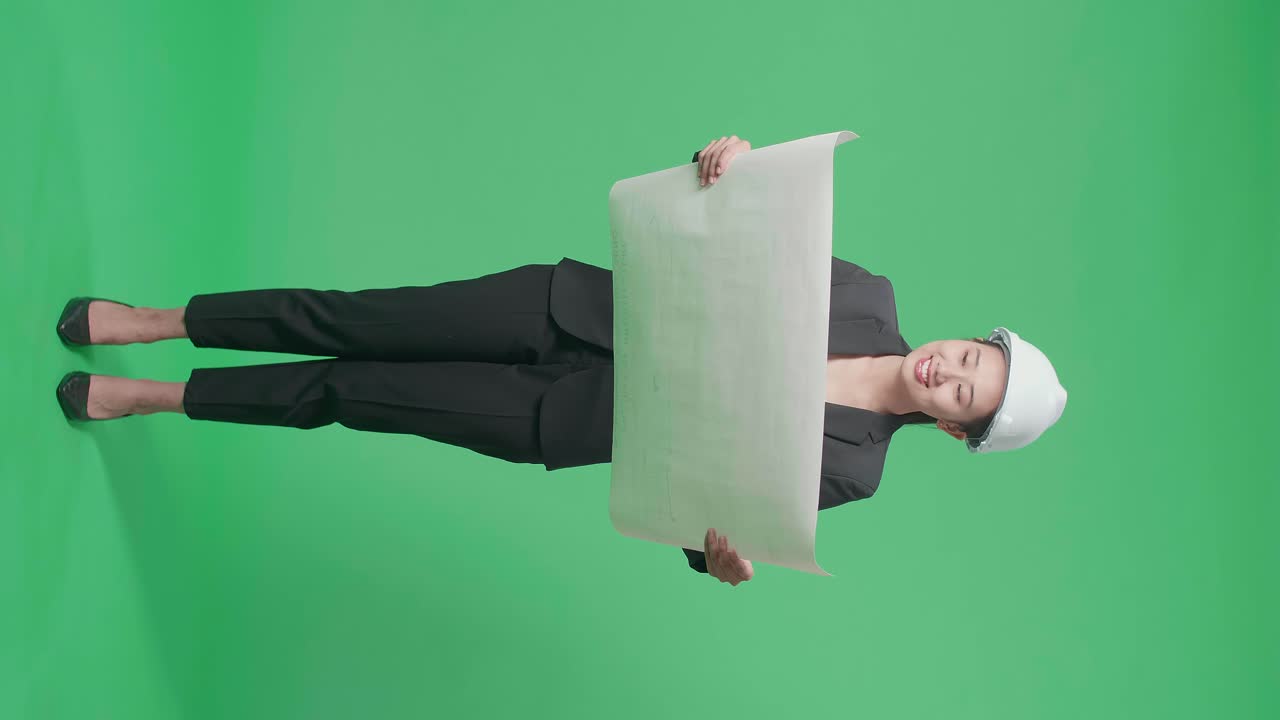 Full Body Of Smiling Asian Female Engineer Looking At Blueprint Before Looking Around And Shaking Her Head In The Green Screen Studio
