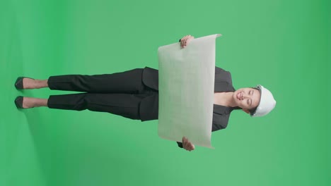 full body of smiling asian female engineer looking at blueprint before looking around and shaking her head in the green screen studio