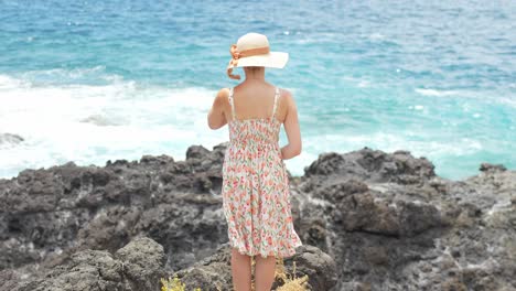 Attractive-woman-in-summer-dress-standing-on-rocky-coastline,-back-view