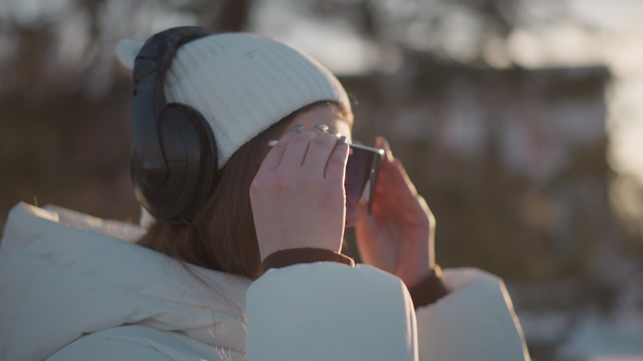 Side view student adjusting tinted goggles under clear sky wearing white beanie and puffer coat as gentle breeze tousles hair with sunshine reflecting off goggles evoking cheerful winter fashion mood