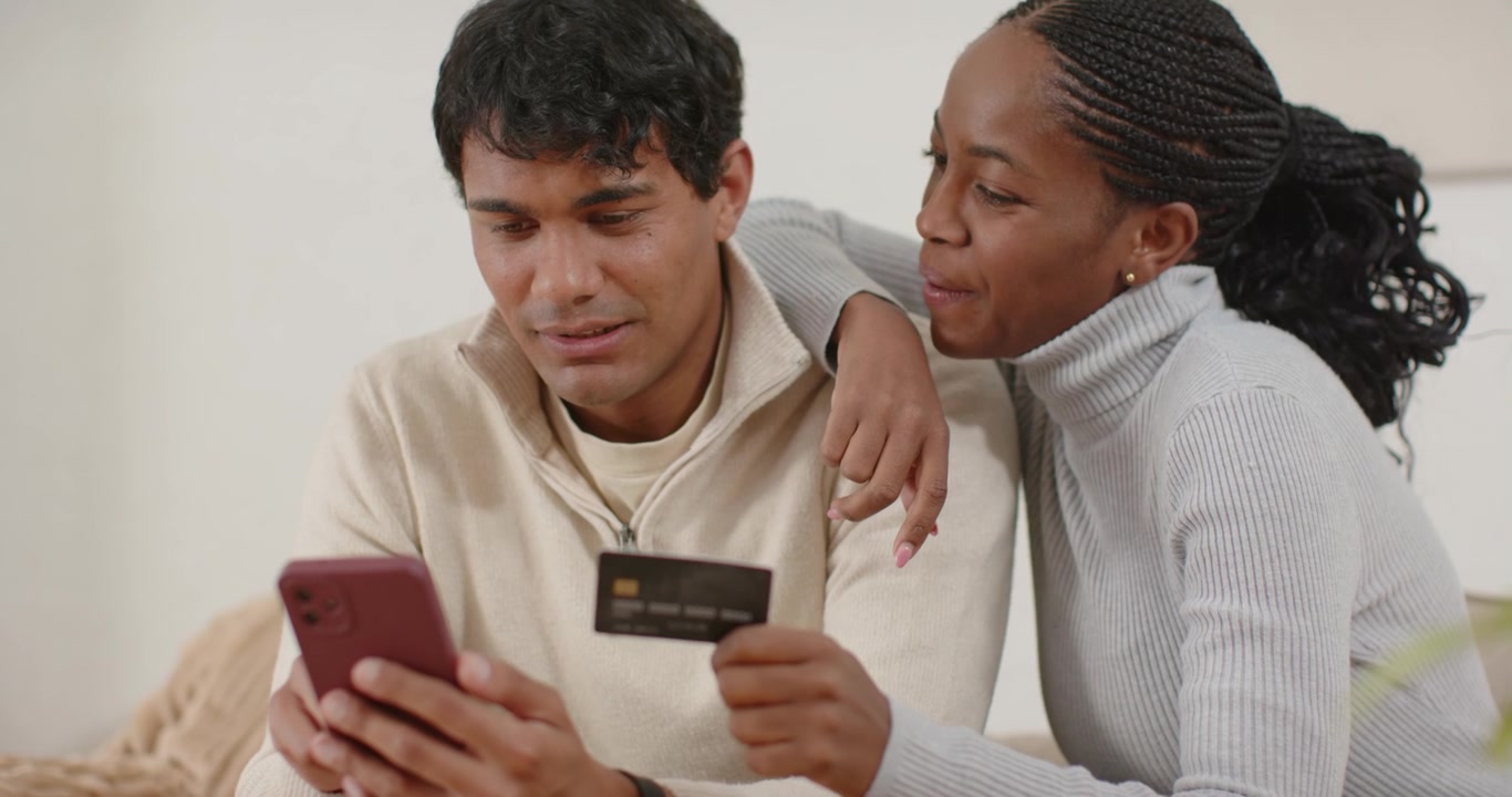 Hispanic man and African American woman confirming purchase on sofa using pink phone and dark card
