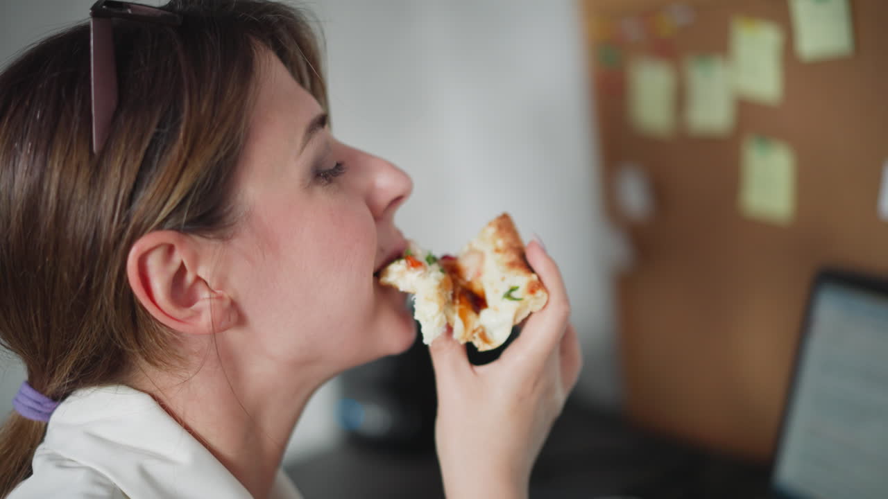 Close up of white lady wearing white jacket enjoying cheesy pizza slice during break from work in cozy office space, focused, holding delicious slice with melted toppings near mouth