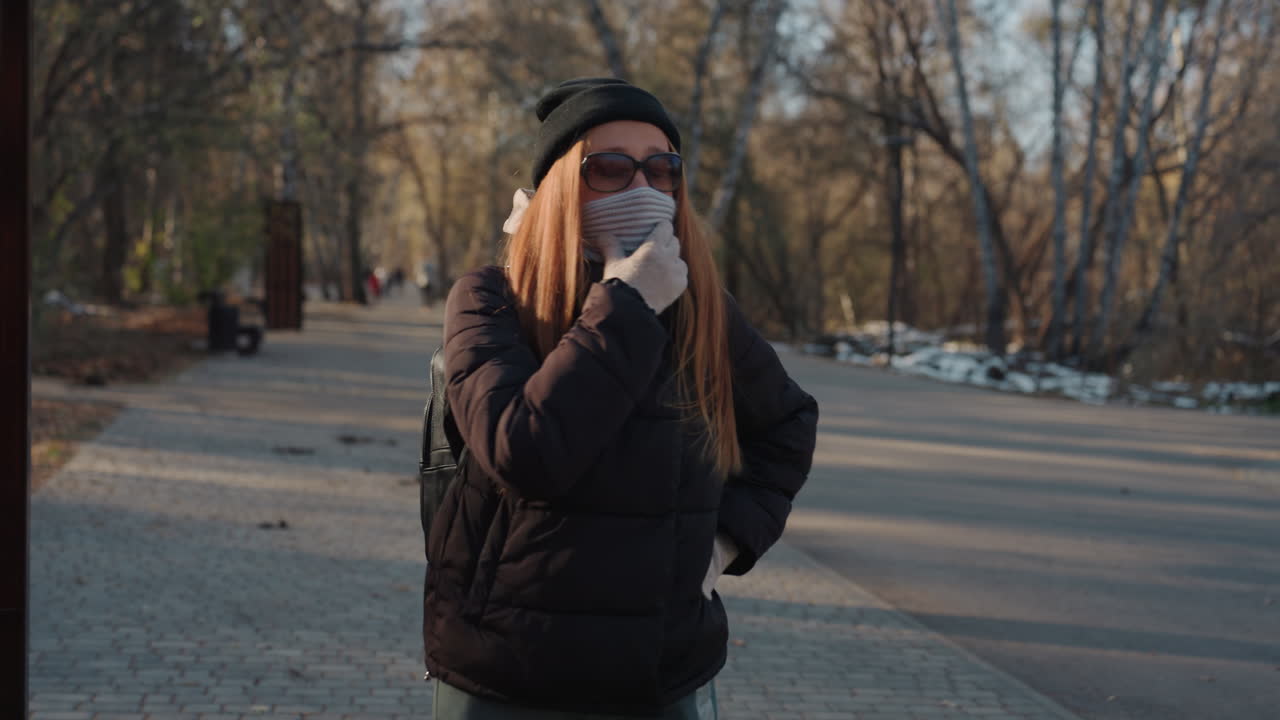 Donna nel parco si copre il viso con sciarpa e occhiali da sole, avvolta in un cappotto scuro e berretto, in piedi su un sentiero autunnale deserto, si guarda intorno con circospezione come se cercasse un'opportunità, sotto un sole tardivo e cinematografico.