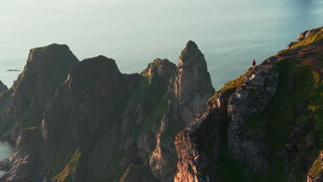 woman on cliff edge by pointed waterfront lofoten mountain in sunset; reveal
