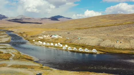 Drones-Aéreos-Que-Recorren-Un-Río-Natural-Con-Lujosas-Tiendas-Blancas-De-Glamping-En-La-Llanura-Alpina-De-Gran-Altitud-Del-Parque-Nacional-Deosai,-Ubicada-Entre-Skardu-Y-El-Valle-De-Astore-En-Pakistán-En-Verano